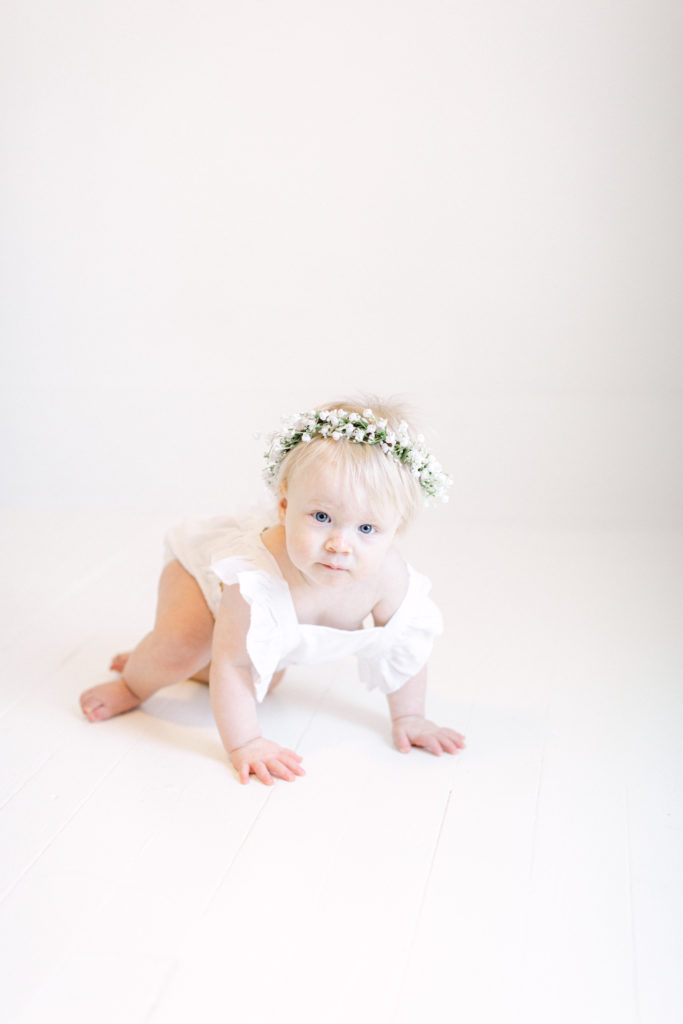 One year old girl sitting on the floor in a white romper with baby's breath flower crown on by Ellen Grace Photography a Cincinnati Family Photographer