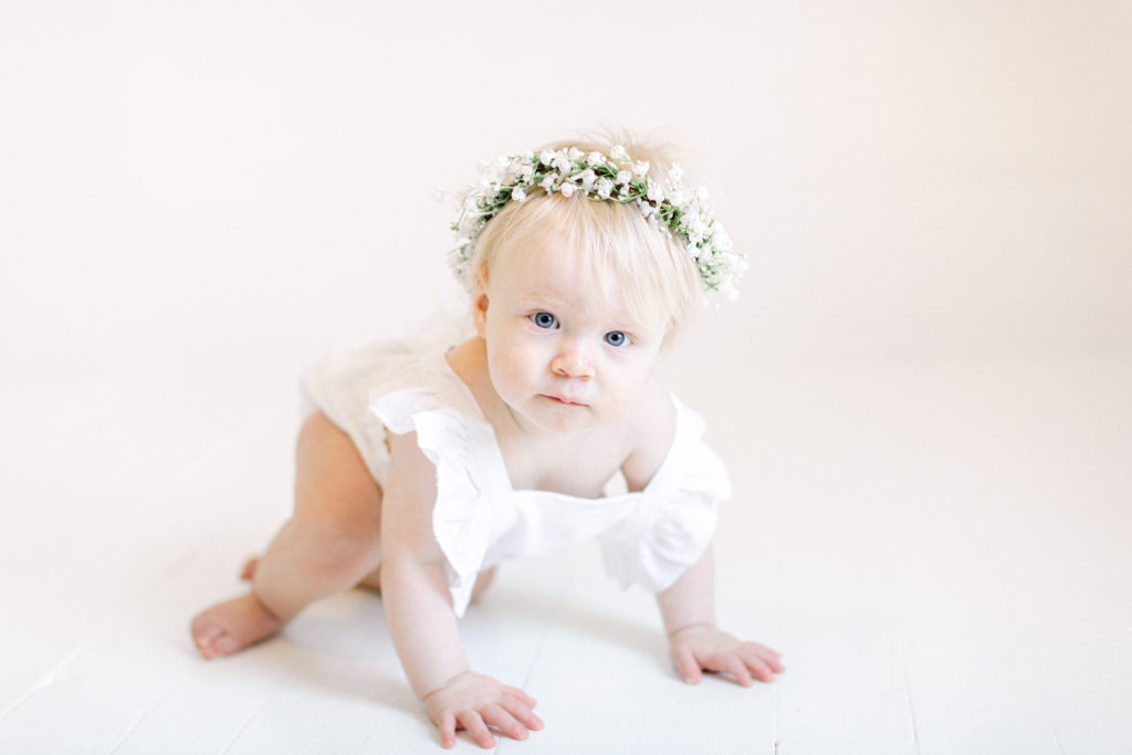One year old girl crawling on the floor in a white romper with baby's breath flower crown on by Ellen Grace Photography a Cincinnati Family Photographer