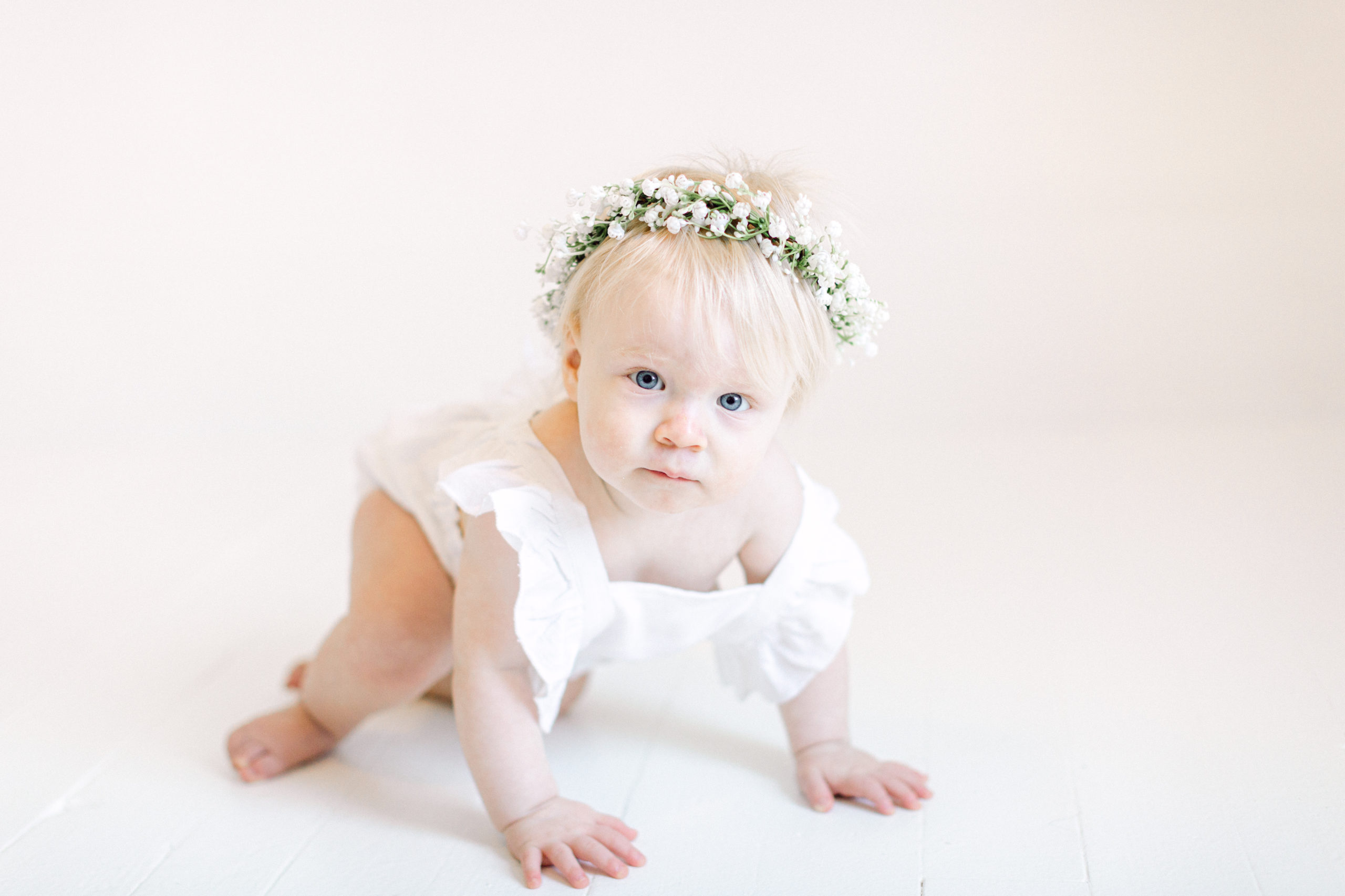 One year old girl crawling in a white romper and flower crown
