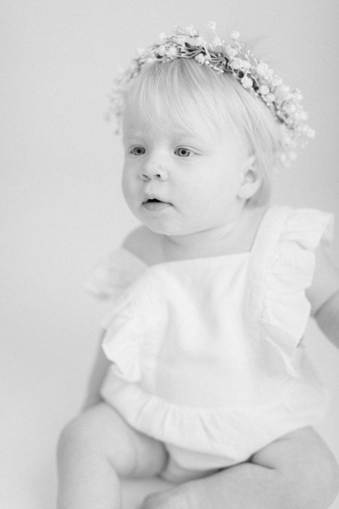 Black and white photo of a one year old girl sitting on the floor in a white romper with baby's breath flower crown on by Ellen Grace Photography a Cincinnati Family Photographer