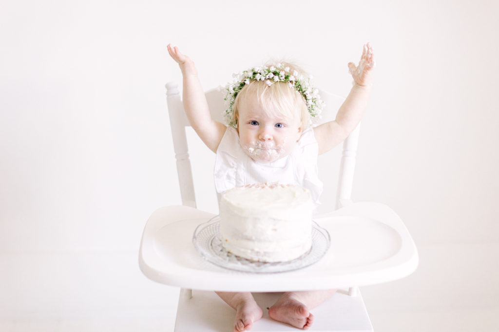 One year old girl in a white romper and flower crown, sitting in a white high chair with a white birthday cake by Ellen Grace Photography a Cincinnati Family Photographer