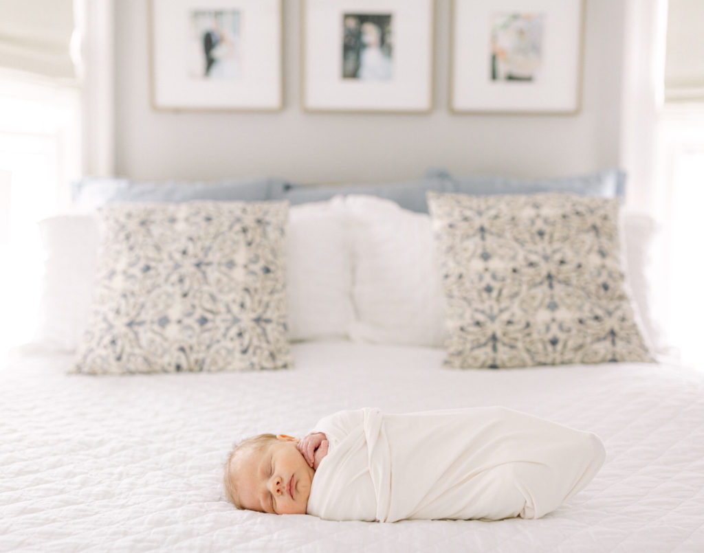 Newborn baby boy swaddled in white blanket laying on his parent's bed next to blue and white pillows taken by Hyde Park Newborn Photographer