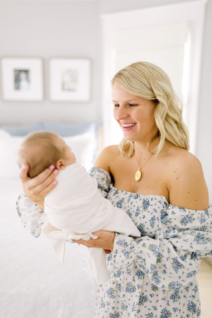 Mother in blue floral dress is smiling as she holds her swaddled newborn son in a white blanket, taken by Hyde Park Newborn Photographer