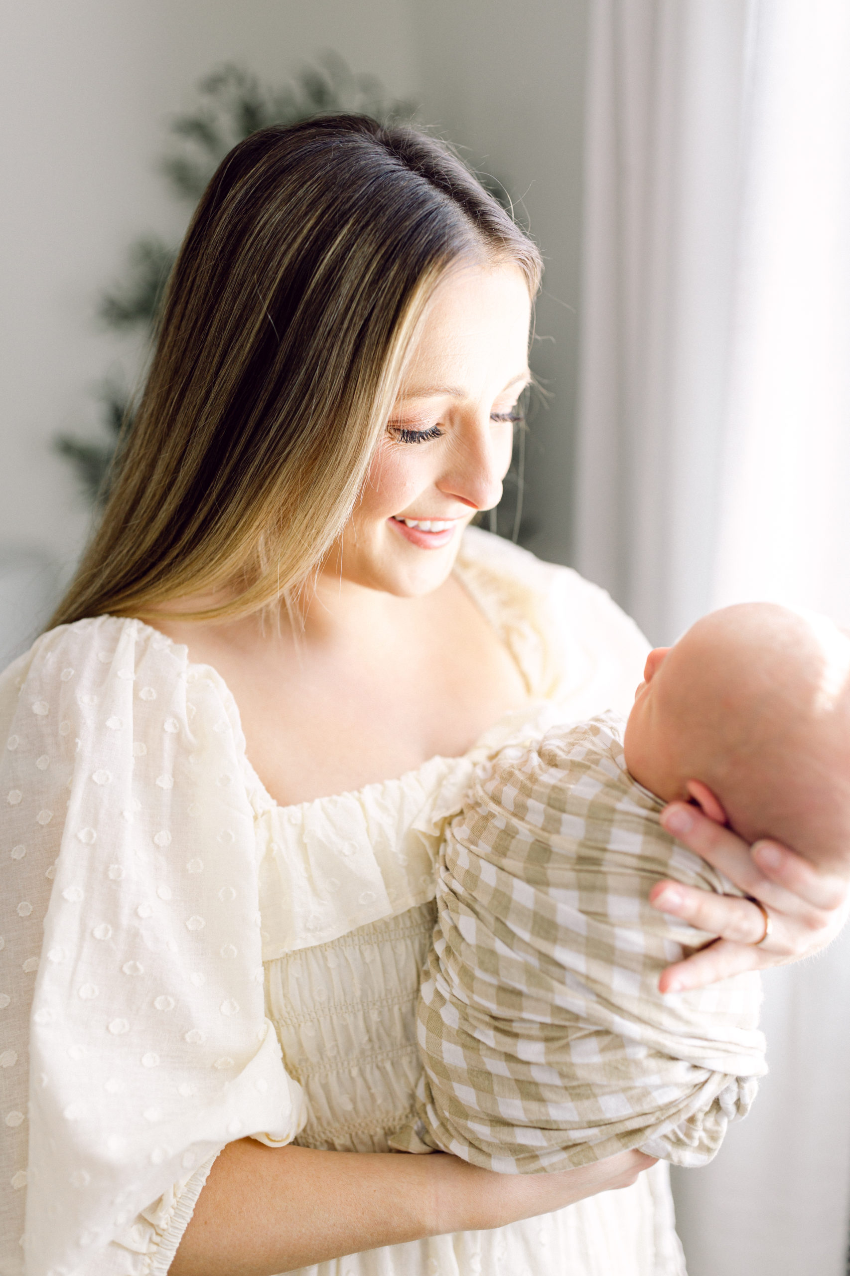 Mother in flowing white dress, smiling down at her swaddled newboen baby taken by Ellen Grace Photography